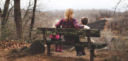 A woman sits between a young boy and a young girl on a bench in a secluded wood. They are facing away from the camera.