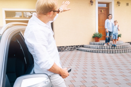 A father waves goodbye to his wife and child while standing beside his car.