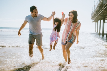 Parents holding hands with child while jumping in water on beach.