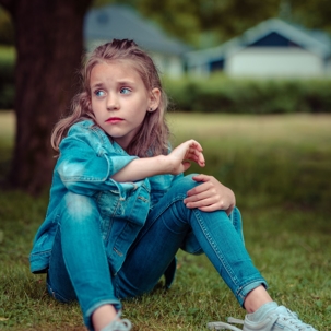 A young girl sits in the grass with one knee in her hand, looking uncertainly to her right.