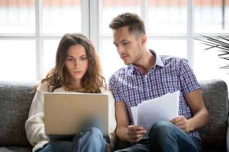 Focused worried couple paying bills online on laptop with documents sitting together on sofa at home