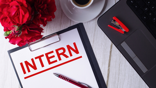 The word INTERN is written in red on a white notepad near a laptop, coffee, red roses and a pen.