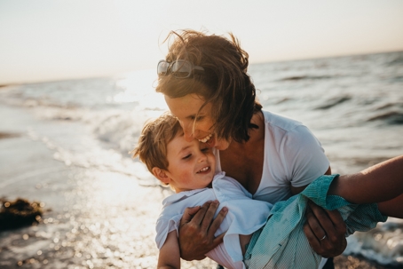 A laughing mother embraces her small child while standing in front of ocean waves