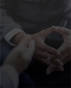 A business person sits with laced fingers while another person speaks to them. Only their hands are shown.