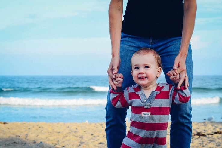 A parent stands on a beach holding the hands of their small child